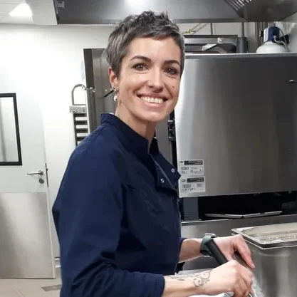 Femme souriante en cuisine, portant un vêtement de chef, préparant un plat.