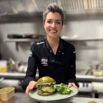 Une cheffe souriante présente un burger sur une assiette avec de la salade.