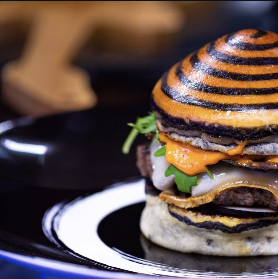Hamburger avec pain rayé noir et orange, viande, fromage et garnitures fraîches.