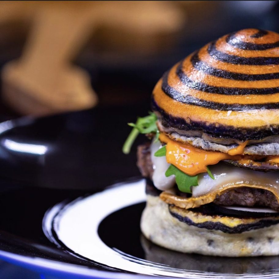 Hamburger avec un pain rayé noir et orange, viande, fromage et garnitures fraîches.